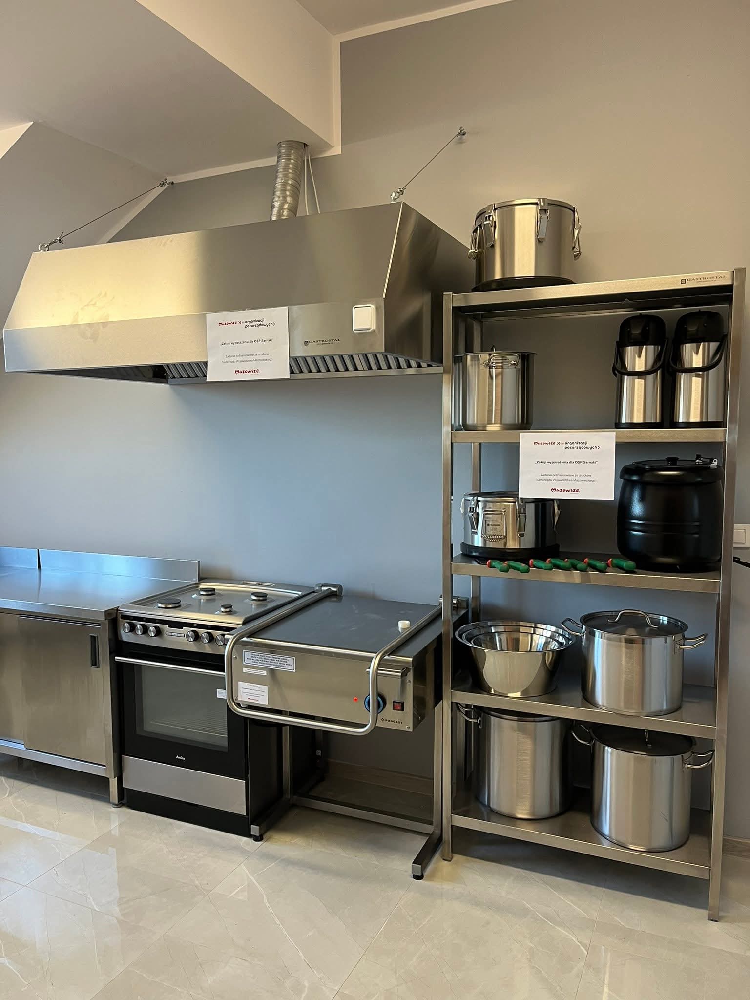 Zaplecze kuchenne z metalowymi urządzeniami: okap wentylacyjny, regał magazynowy, a na nim stoją garnki, termosy gastronomiczne, miski ze stali nierdzewnej, komplet noży i kociołek elektryczny. Pomieszczenie ma jasne ściany i błyszczącą podłogę.