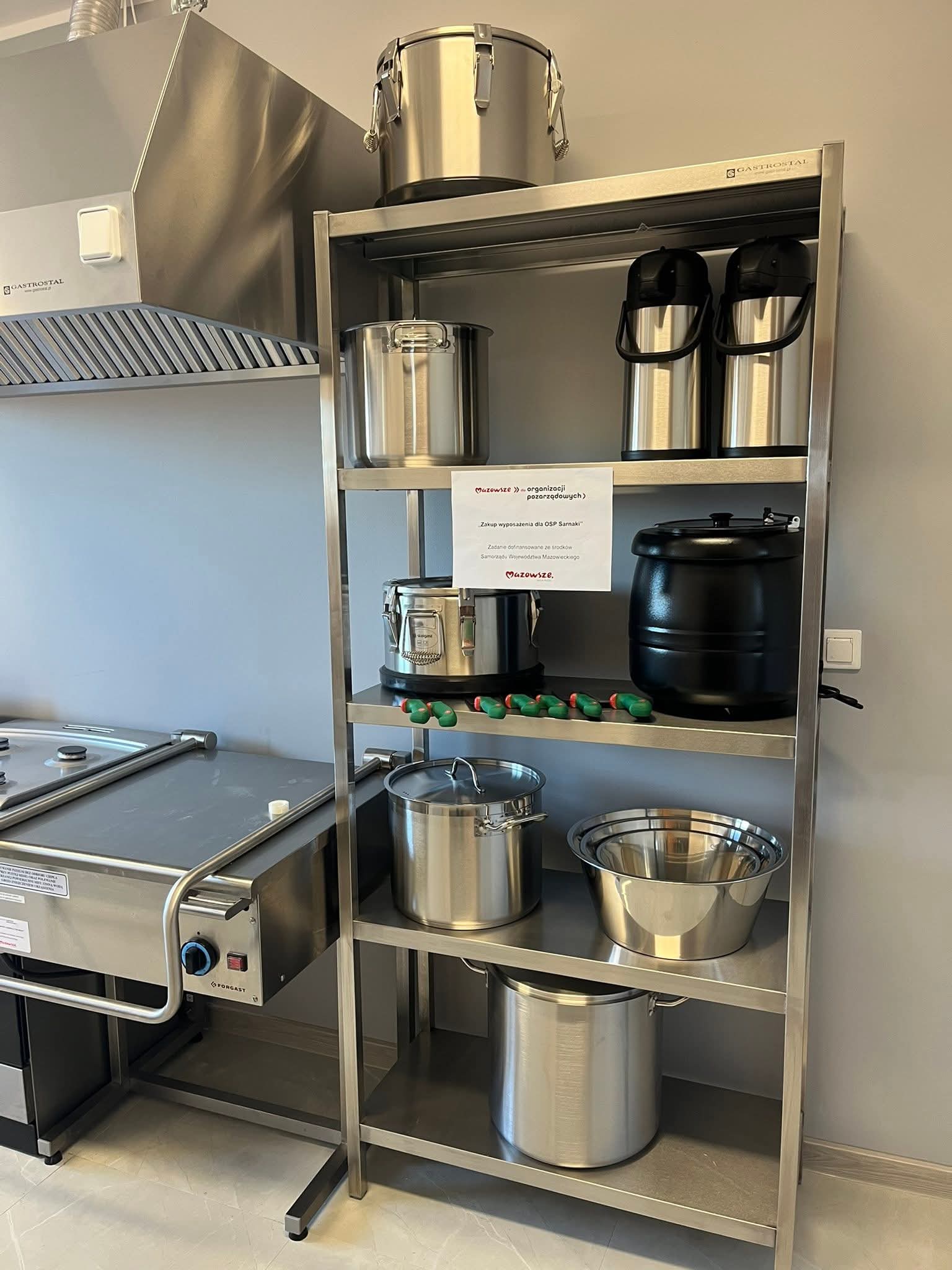 Zaplecze kuchenne z metalowymi urządzeniami: okap wentylacyjny, regał magazynowy, a na nim stoją garnki, termosy gastronomiczne, miski ze stali nierdzewnej, komplet noży i kociołek elektryczny. Pomieszczenie ma jasne ściany i błyszczącą podłogę.