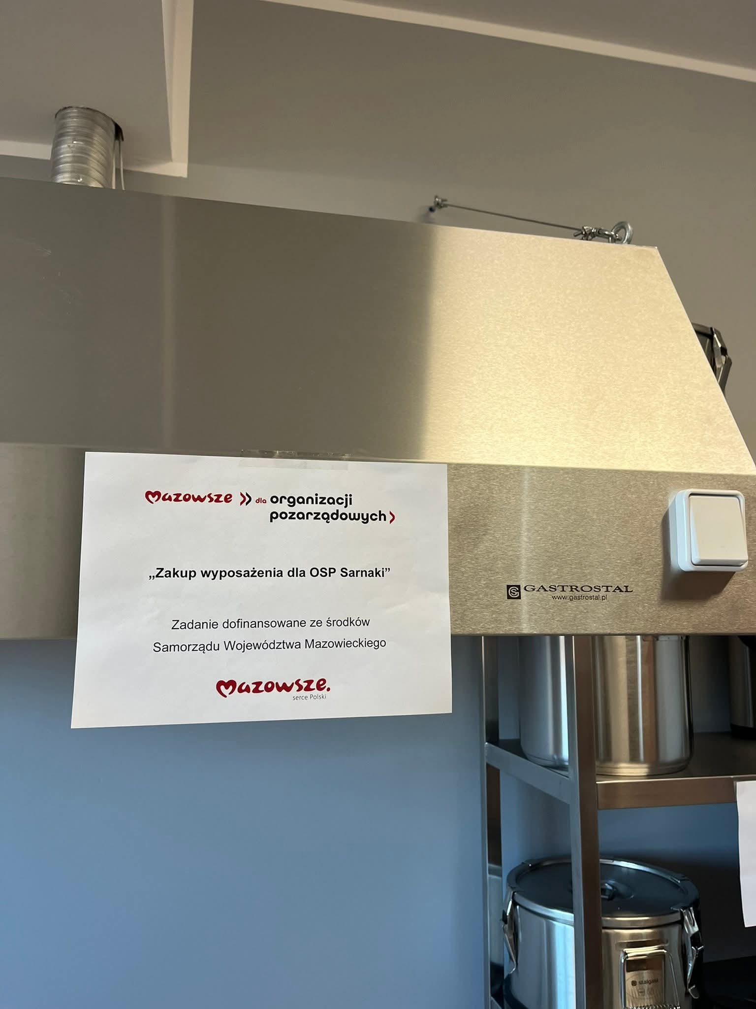 Zaplecze kuchenne z metalowymi urządzeniami: okap wentylacyjny, regał magazynowy, a na nim stoją garnki, termosy gastronomiczne, miski ze stali nierdzewnej, komplet noży i kociołek elektryczny. Pomieszczenie ma jasne ściany i błyszczącą podłogę.